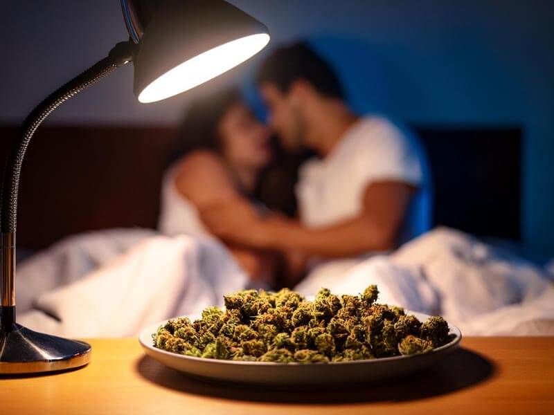 plate of cannabis buds under a lit lamp in a bedroom. Backroom is dimly lit with a couple getting romantic in bed